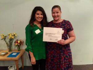 There is a woman with dark brown hair and green shirt stands next to another woman with auburn hair. She is wearing a dress and is holding a certificate of volunteer excellence in her hand