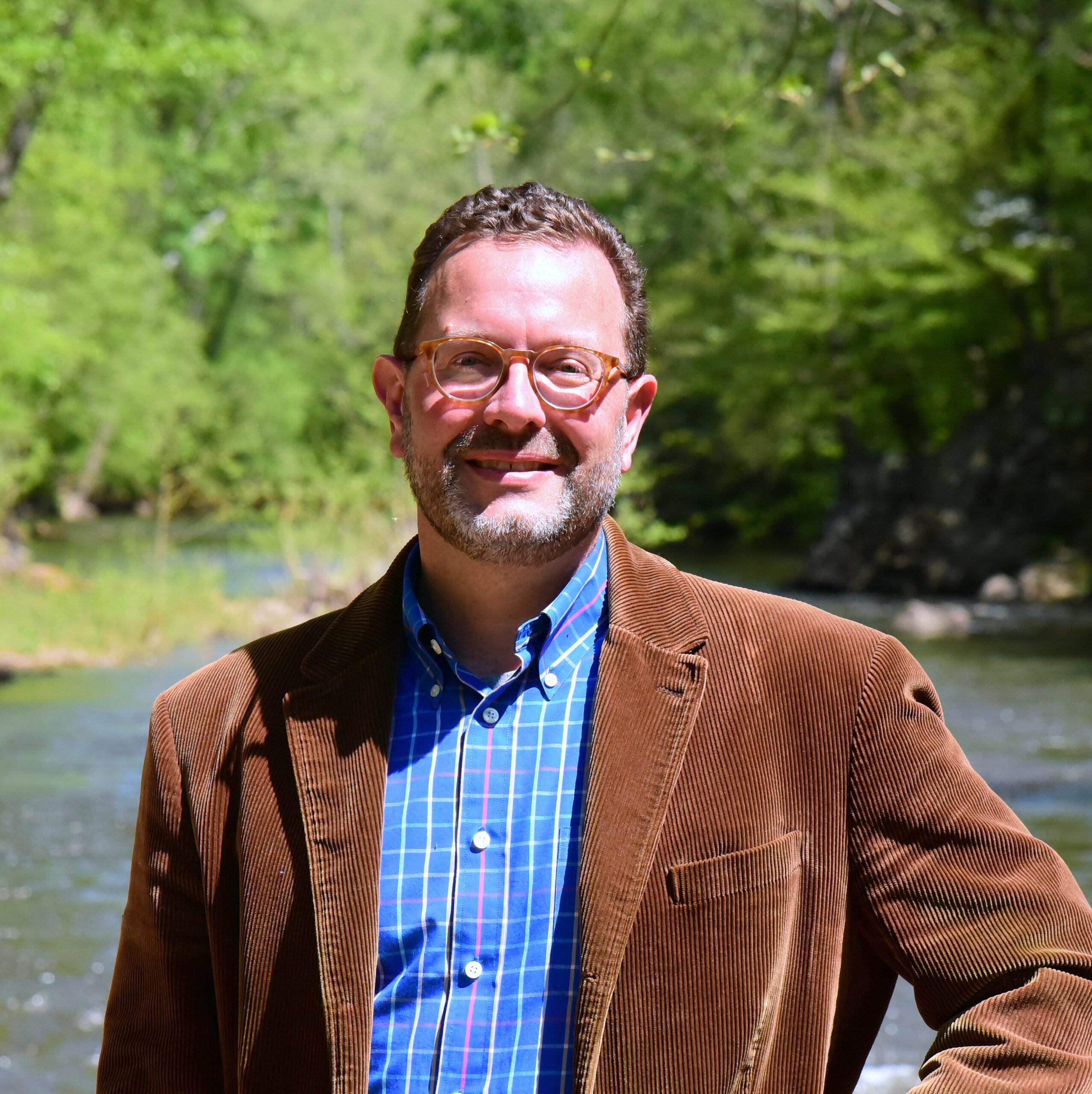 Ryan Fehrman headshot, wearing a brown blazer over a blue button-down shirt. Eno River in the background.